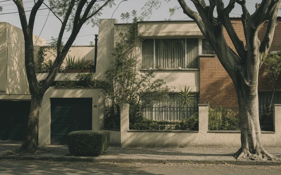 Casa de dos plantas de los años sesenta en una calle arbolada: azotea plana, aplanado pintado, barandal de herrería y puerta de cochera a pie de calle.